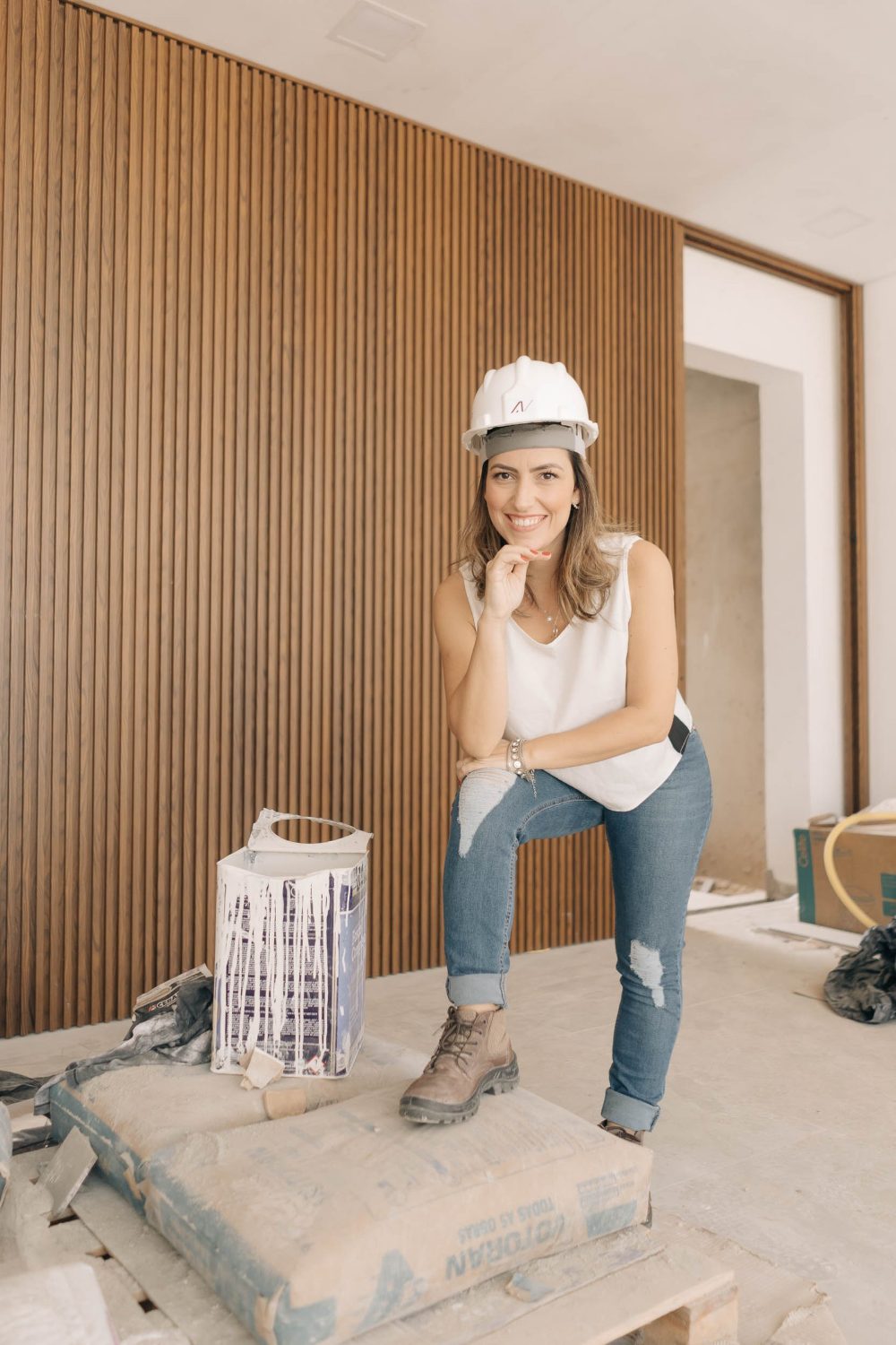 Nayara Barros de capacete na obra, ao lado de um painel de madeira ripada